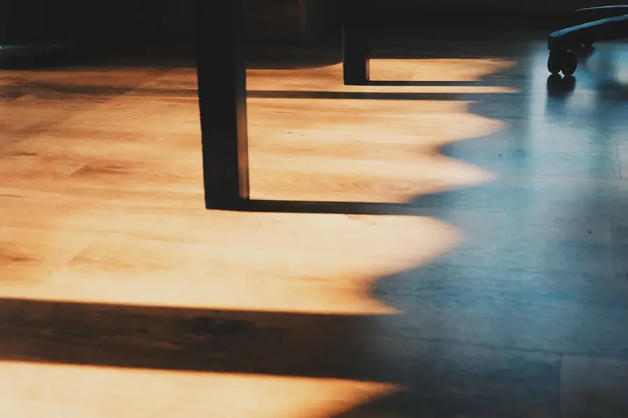 Blick auf einen Parkett Holzboden mit starkem, warmem Lichteinfall. Eine markante, wellenförmige Schattengrenze teilt die Fläche. Im Schatten stehen zwei schwarze, eckige Tischbeine und am rechten Rand die Rollen eines Bürostuhls.